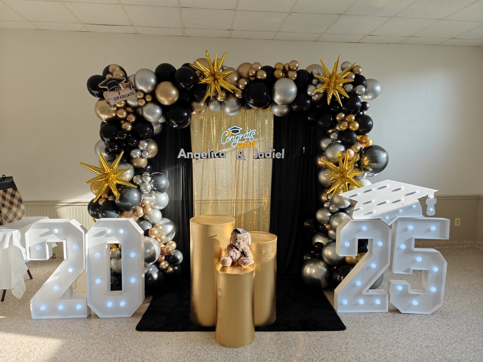 Graduation decor with black, gold, and silver balloon arch, marquee numbers, metallic stars, and custom sign
