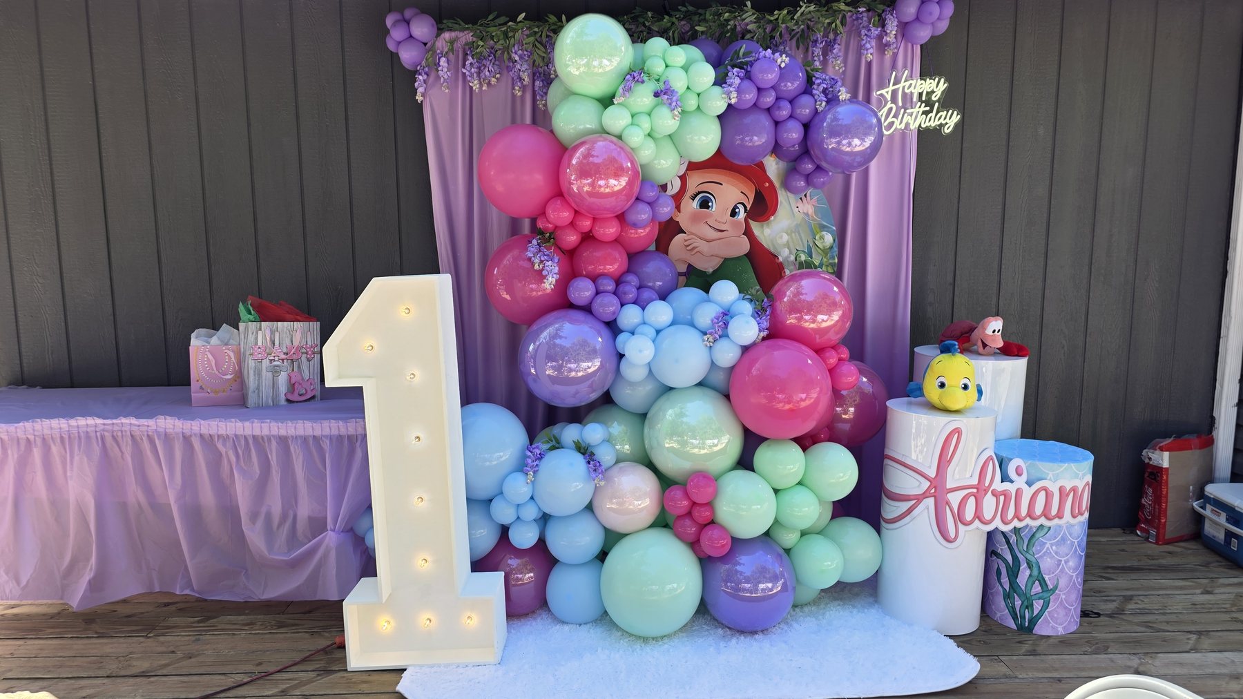 Pastel mermaid first birthday decor with lavender draping, balloons, marquee number, and custom cylinders